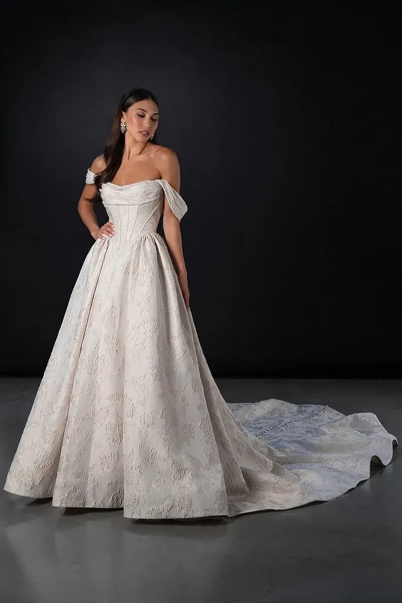 Model is wearing a silk ballgown with detachable off-the-shoulder straps, a scooped neckline, tailored corset bodice with Basque waist, and a buttoned long train.