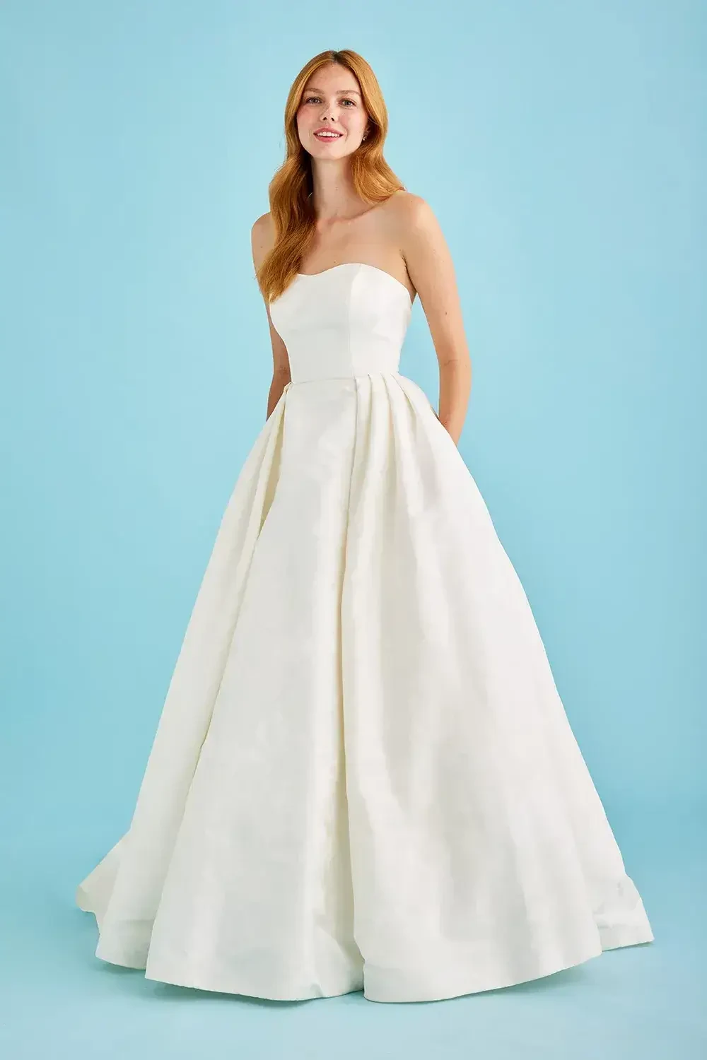 A woman in a strapless, white ball gown stands against a light blue background. She has long, reddish hair and a confident, smiling expression.