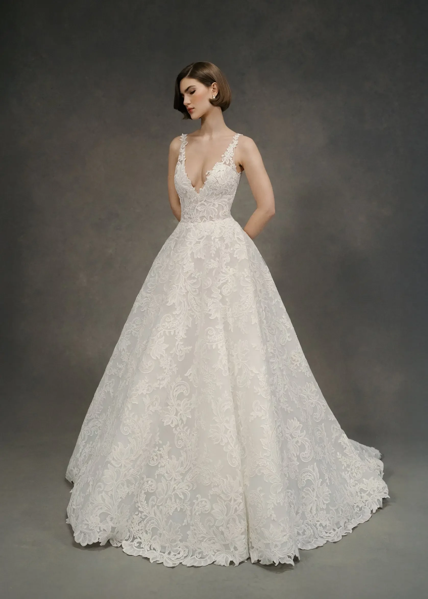 A woman in an elegant white lace wedding gown with a deep V-neckline poses against a dark, neutral background. Her expression is calm and serene.