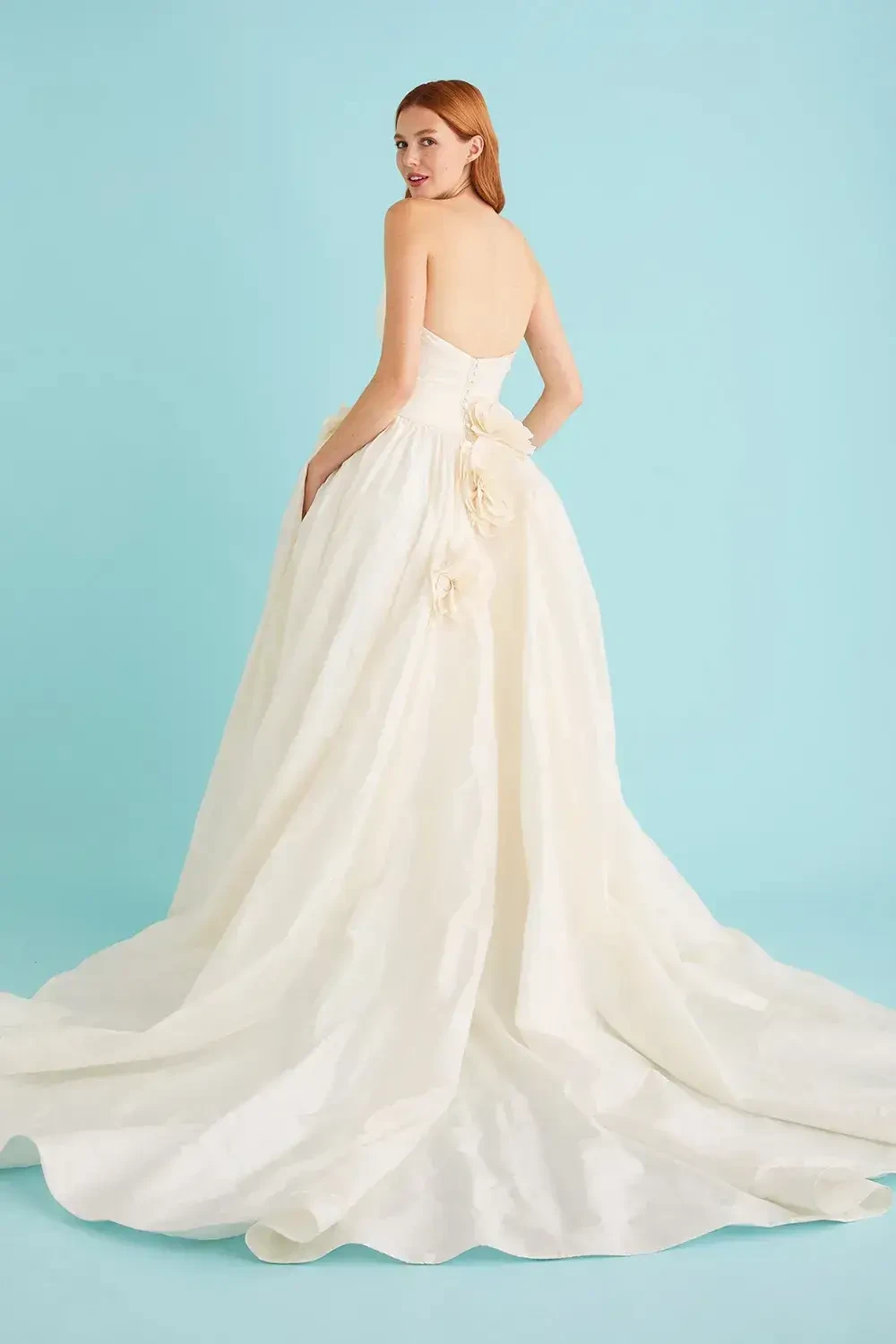 Model is wearing a strapless taffeta ball gown with ruching on the bodice, hand-cut peony appliqués at the hip and low back, and a full gathered skirt with chapel train.