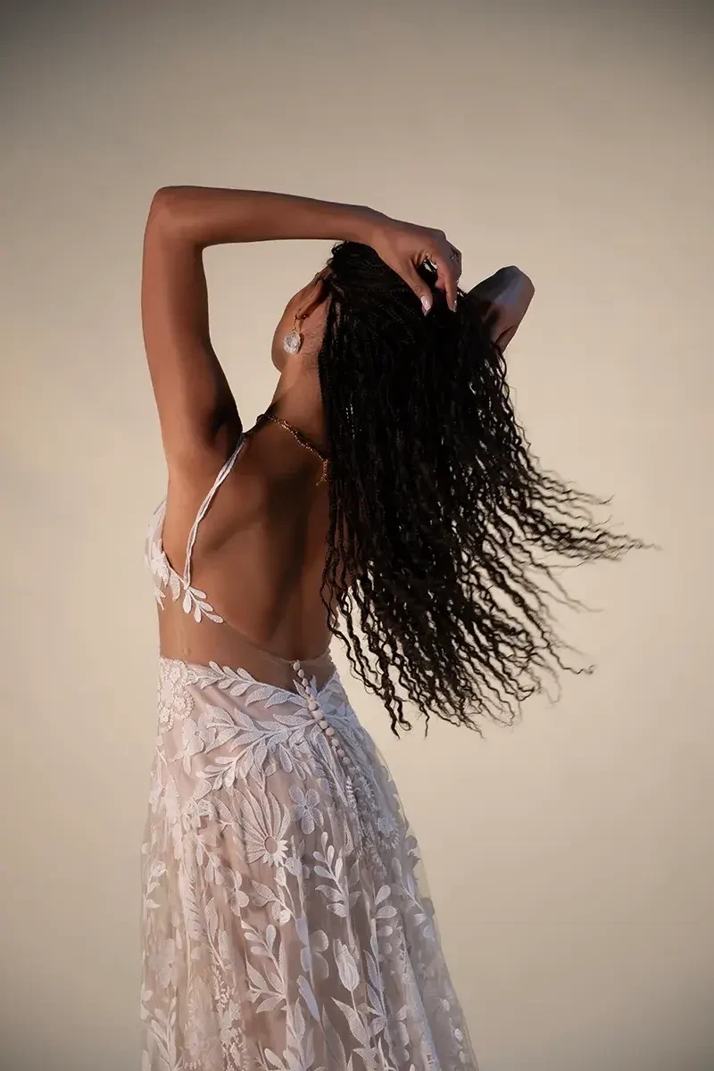 A woman in a flowing floral dress stretches her arms back, tilting her head, her long braided hair cascading down gracefully against a neutral backdrop.