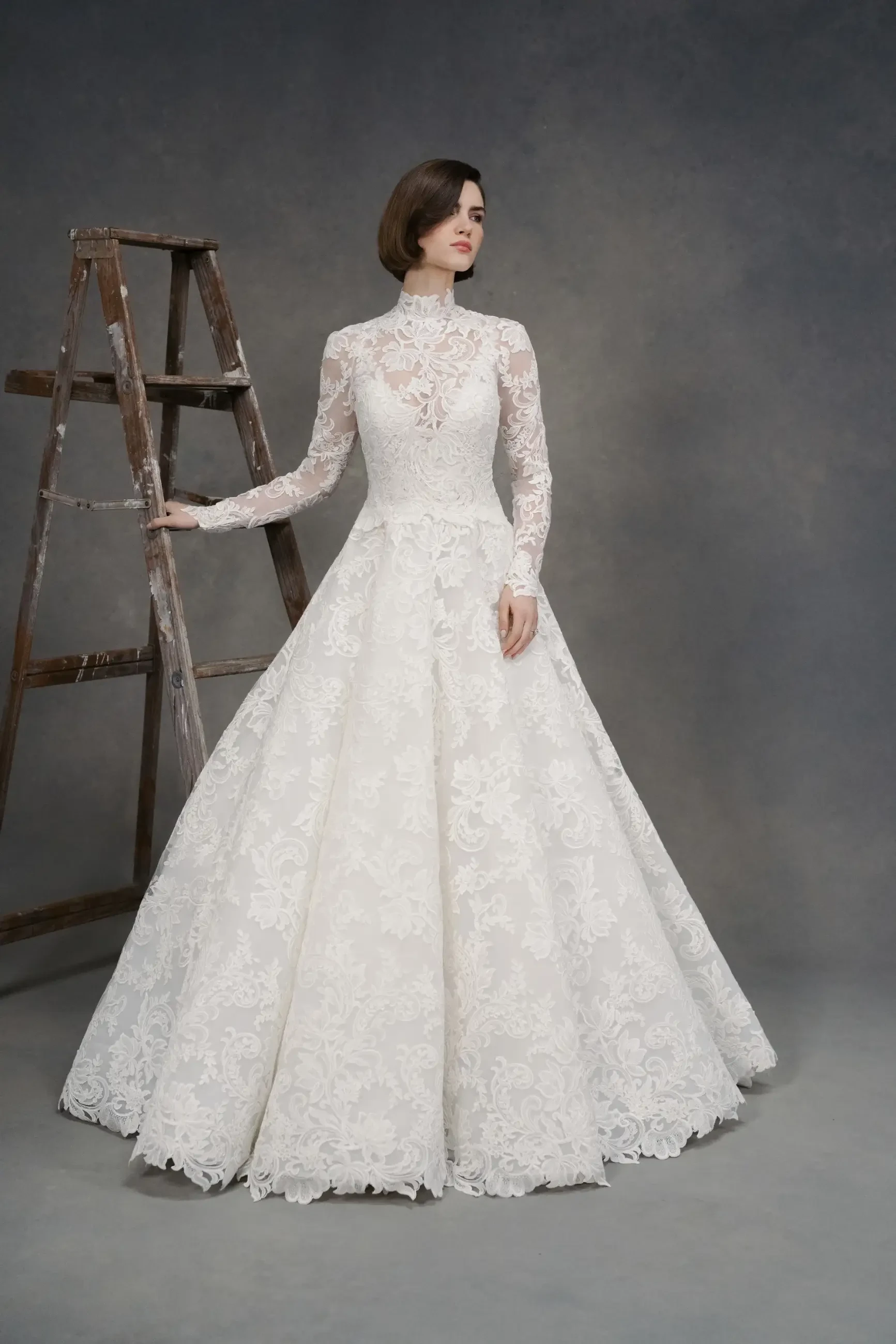 A woman in a vintage white lace wedding dress with a high neck and long sleeves stands near a wooden ladder, set against a muted gray background.