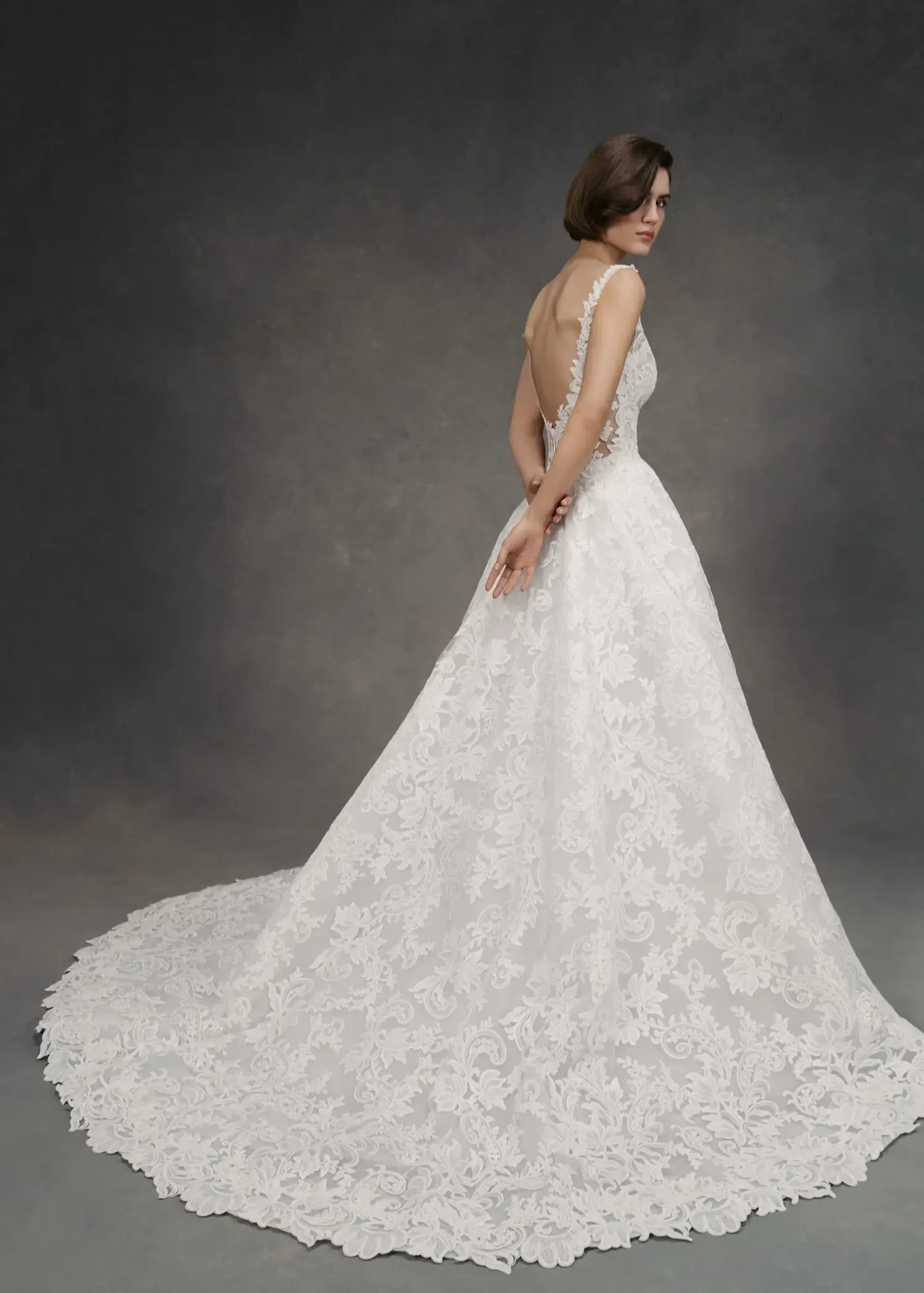 A woman stands elegantly in a white lace wedding dress with a long flowing train. The dress features a low back and floral patterns, evoking a timeless, graceful mood.