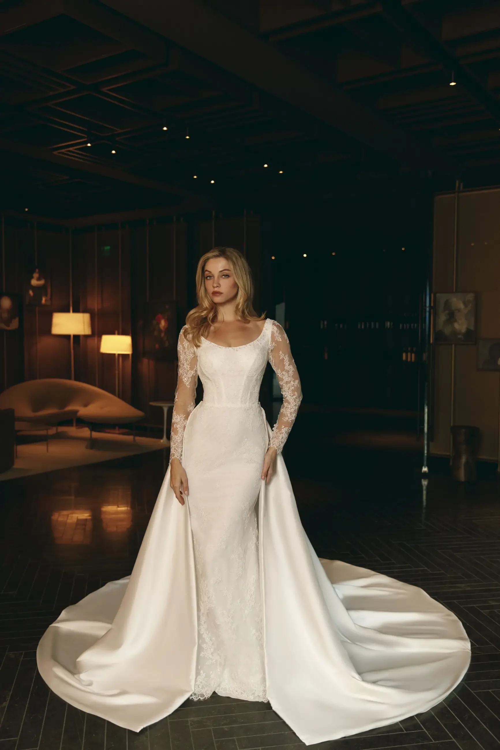 A woman in an elegant, long-sleeved, white wedding gown with a flowing train stands in a dimly lit, sophisticated room, exuding a timeless elegance.