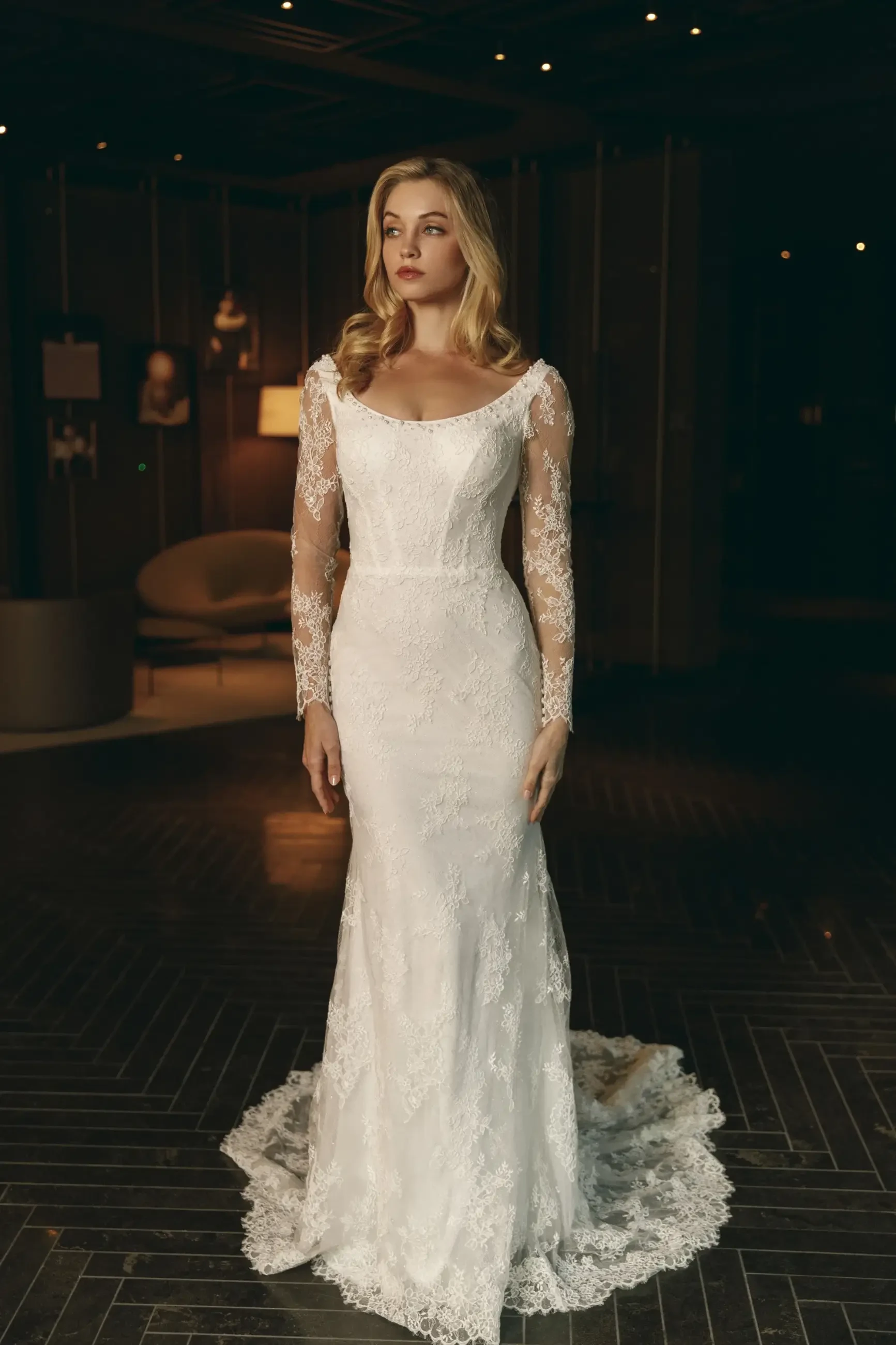 A woman stands elegantly in a dimly lit room wearing a lace wedding gown with long sleeves and a train. The mood is sophisticated and serene.