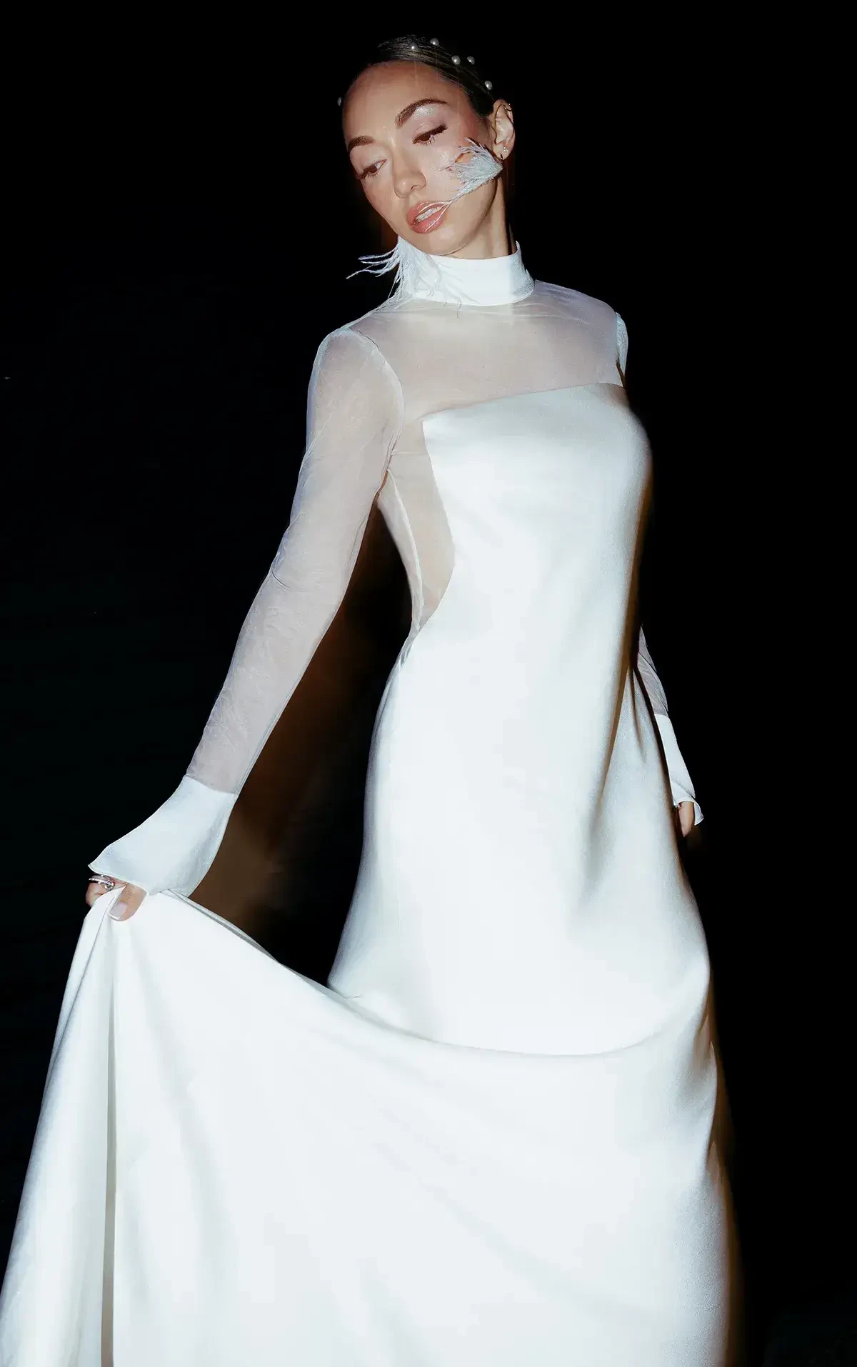 Woman in an elegant, long-sleeved white gown stands gracefully against a dark background. Her serene expression conveys a sense of calm and sophistication.