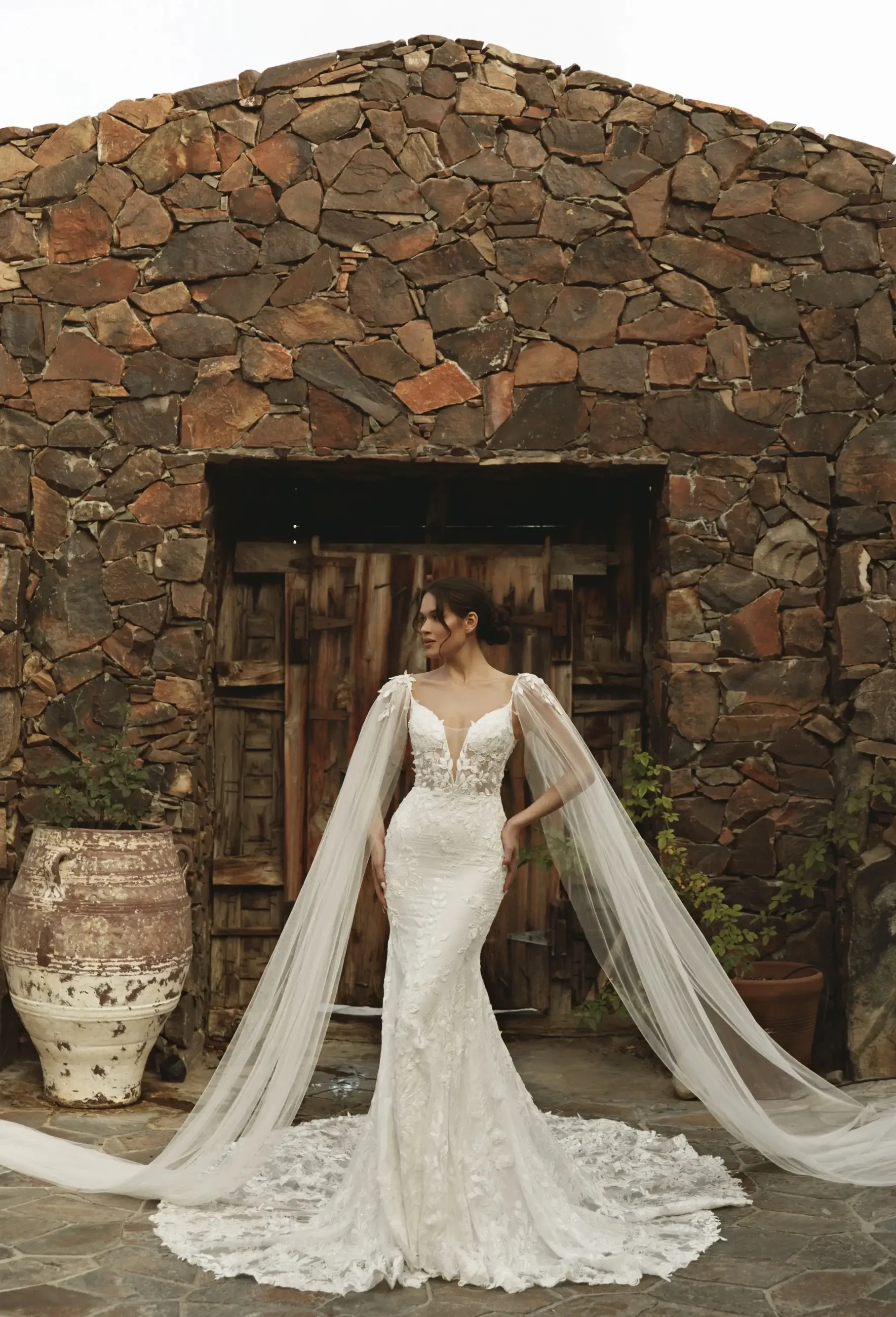 A woman in a stunning white wedding dress with intricate lace detailing and long sheer sleeves stands gracefully in front of a rustic stone building. The dress has a form-fitting silhouette that flares at the bottom, and she is complemented by flowing fabric trailing behind her.
