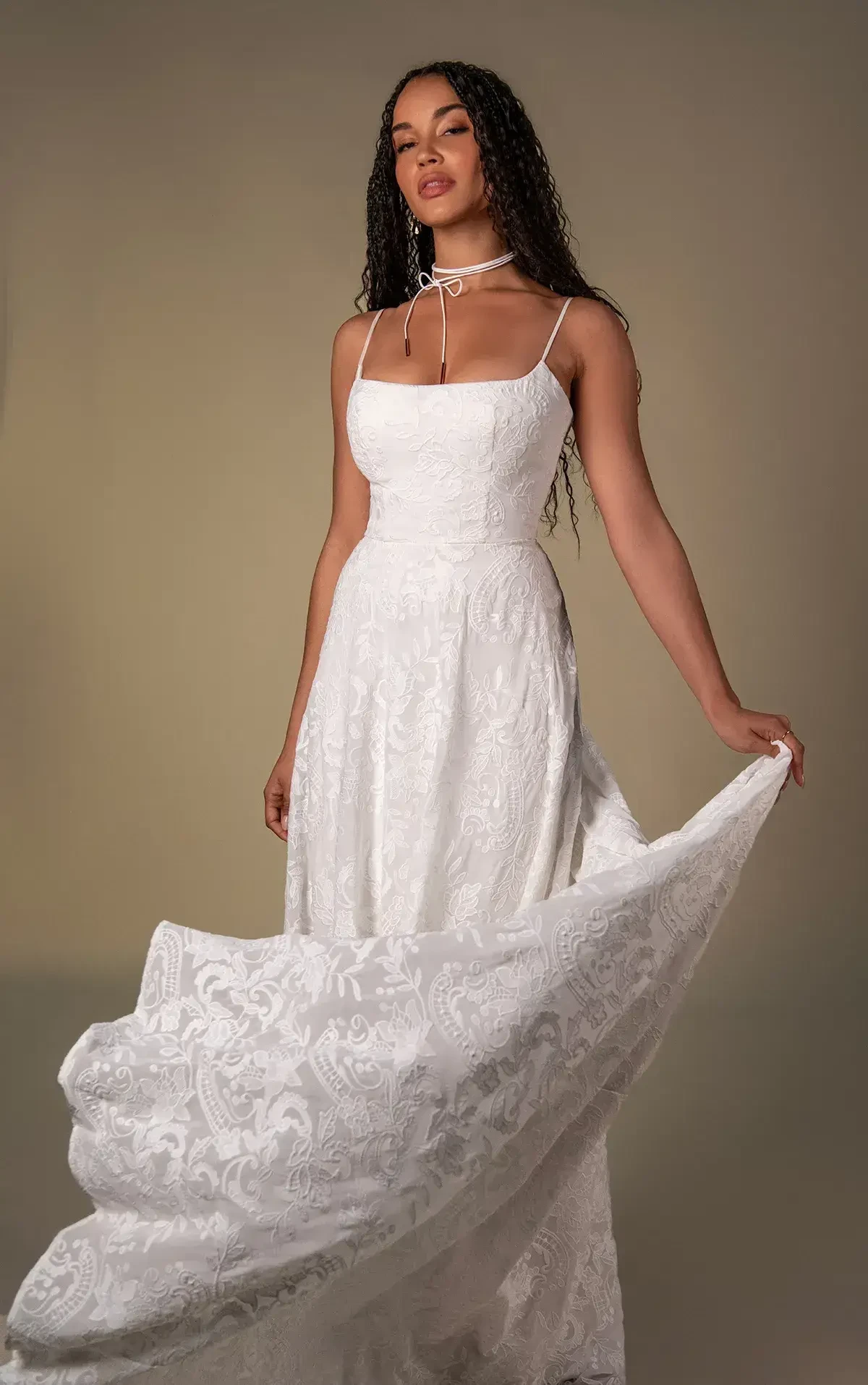 A woman in a white, elegant dress with a fitted bodice and flowing skirt, posing gracefully while holding part of the skirt. She has long, wavy hair and wears a simple choker necklace.