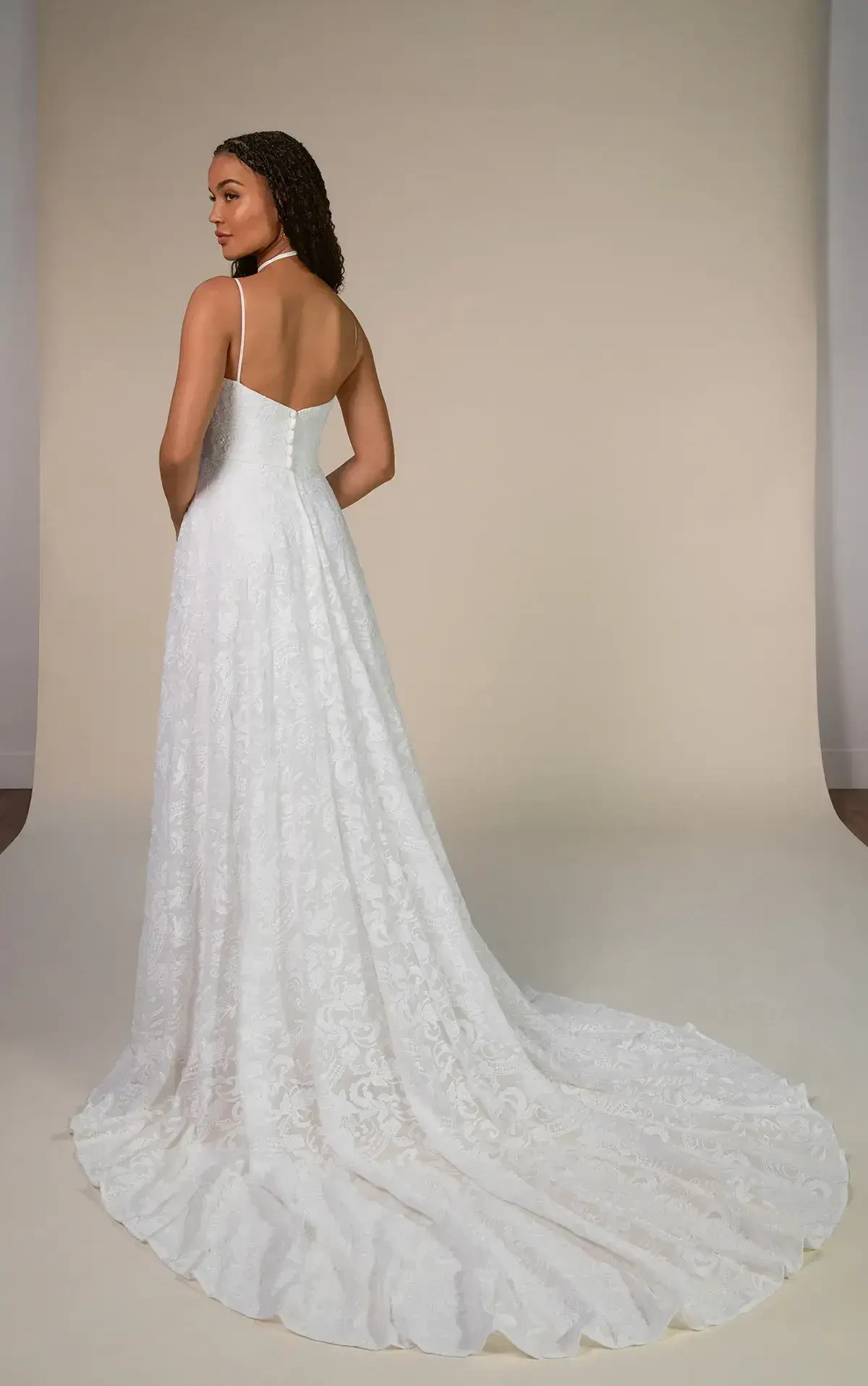 A woman in an elegant white lace wedding gown stands with her back to the camera. The dress features thin straps and a long train. The setting is minimal and serene.