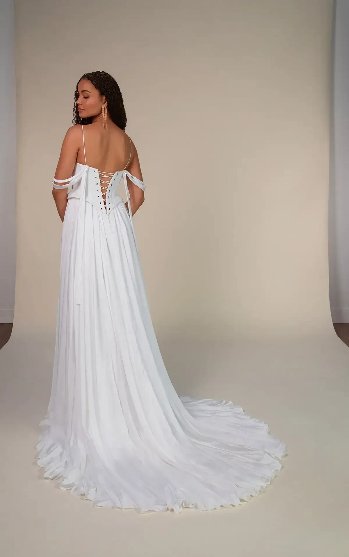 A woman in an elegant white off-shoulder gown stands with her back to the camera. The dress features lace-up details and a long flowing train, conveying grace and sophistication.