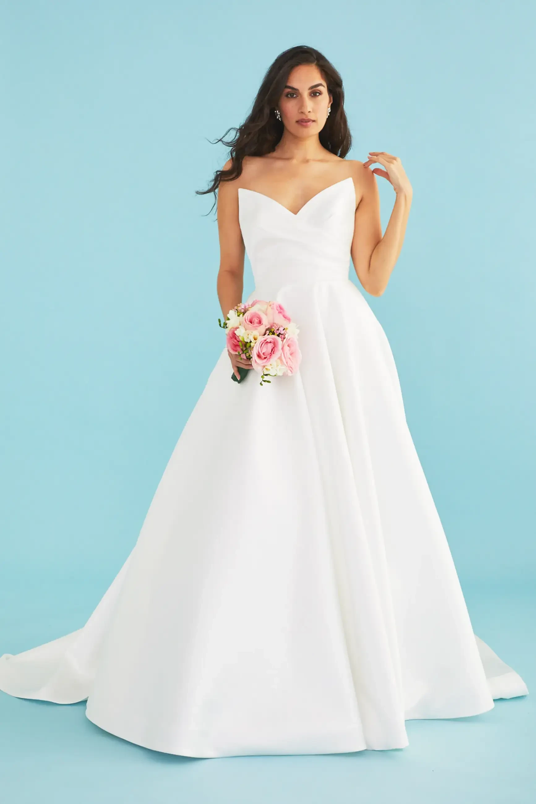A woman in an elegant white strapless wedding gown holds a bouquet of pastel pink flowers. She stands against a light blue background, exuding grace.