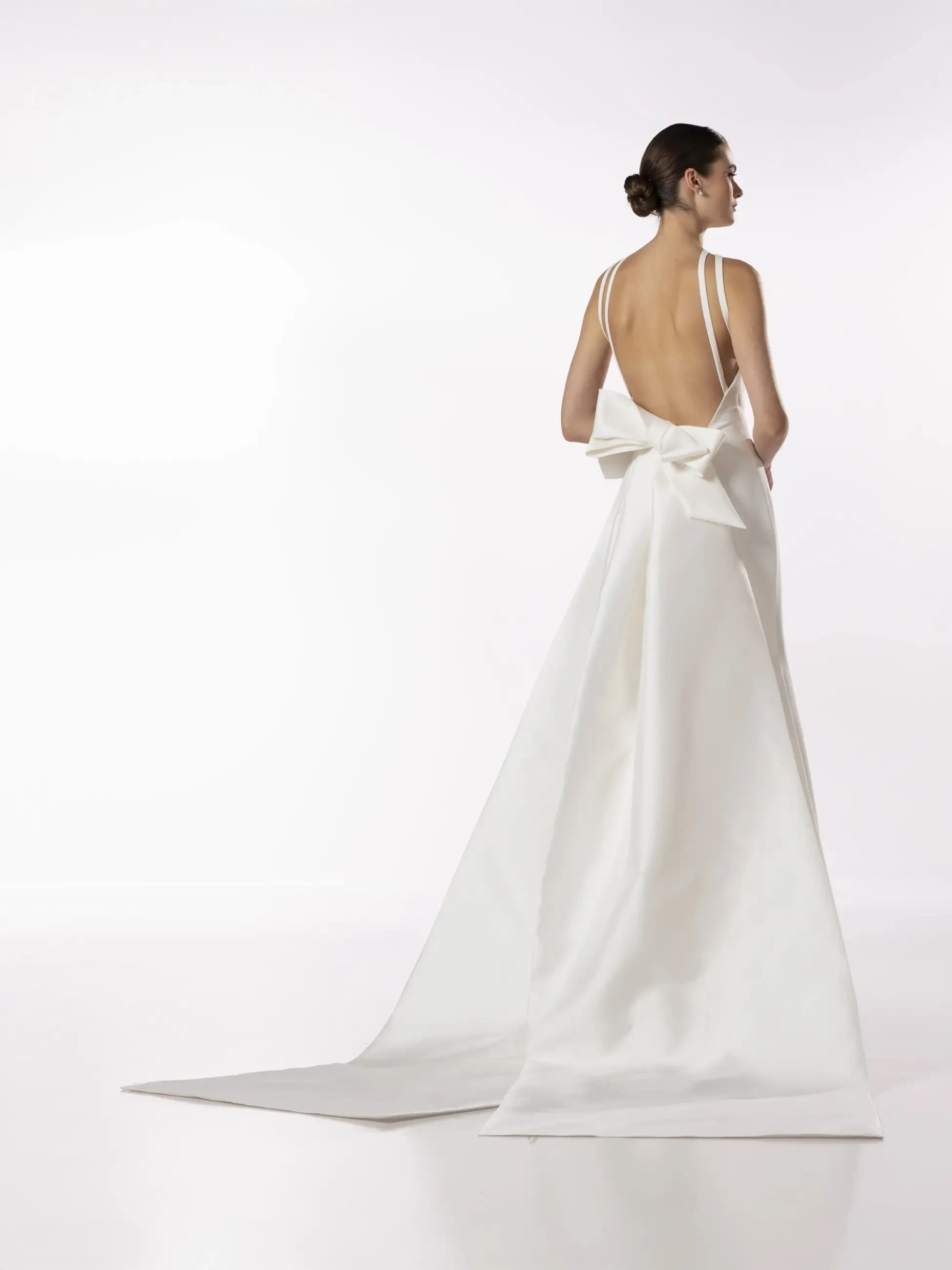 A woman in an elegant white gown with a long train and large bow stands against a minimalistic white background, conveying grace and sophistication.