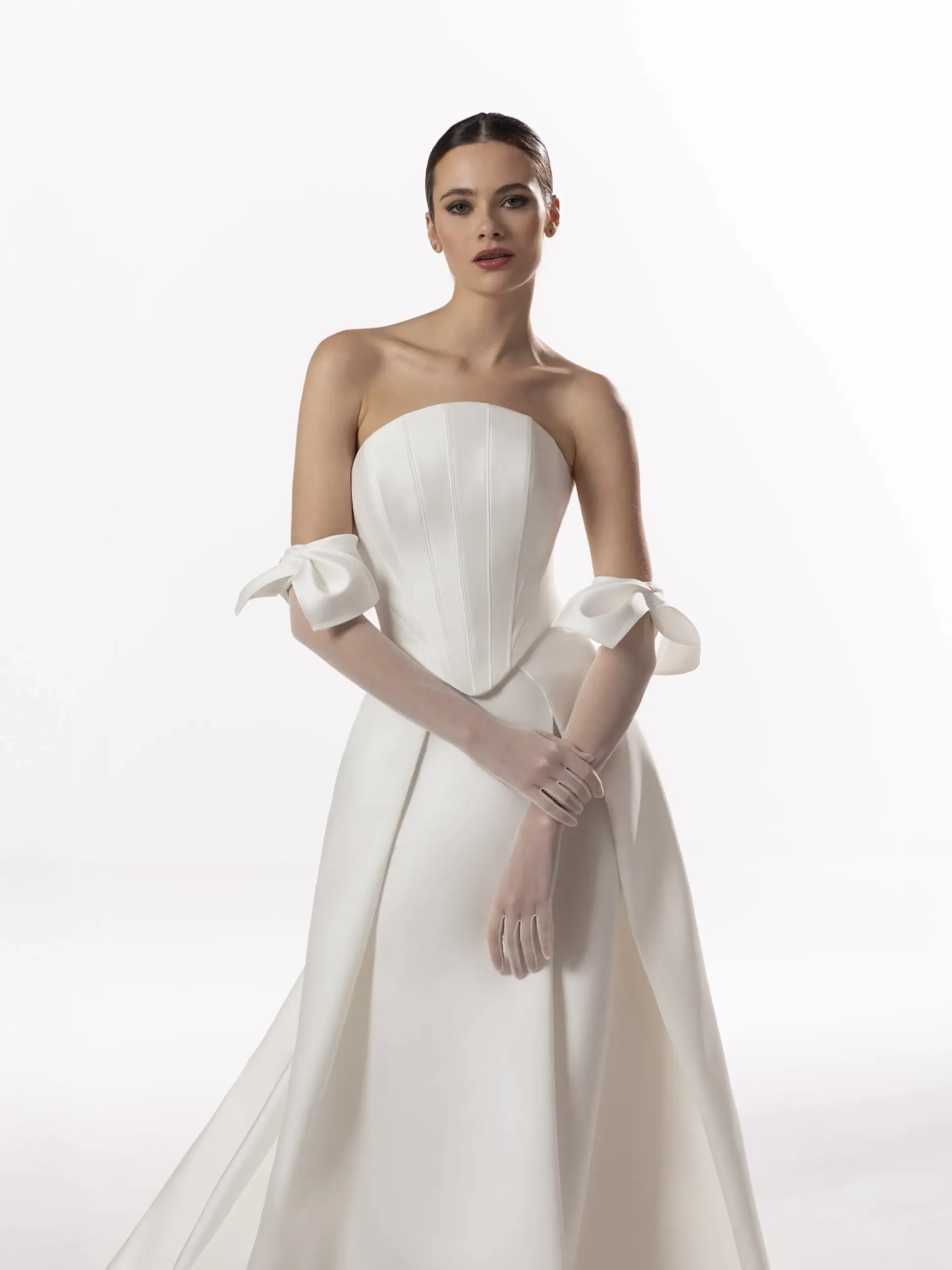 A woman in a strapless white gown with bow details on the sleeves and elegant gloves, standing against a plain background.