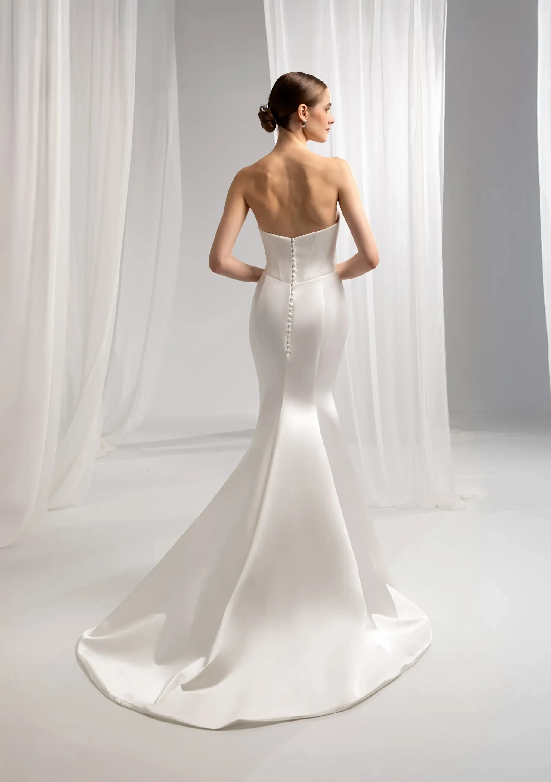 A woman wearing a sleek, strapless white wedding gown, seen from the back, with buttons running down the spine and a train flowing behind. Soft fabric drapes in the background.