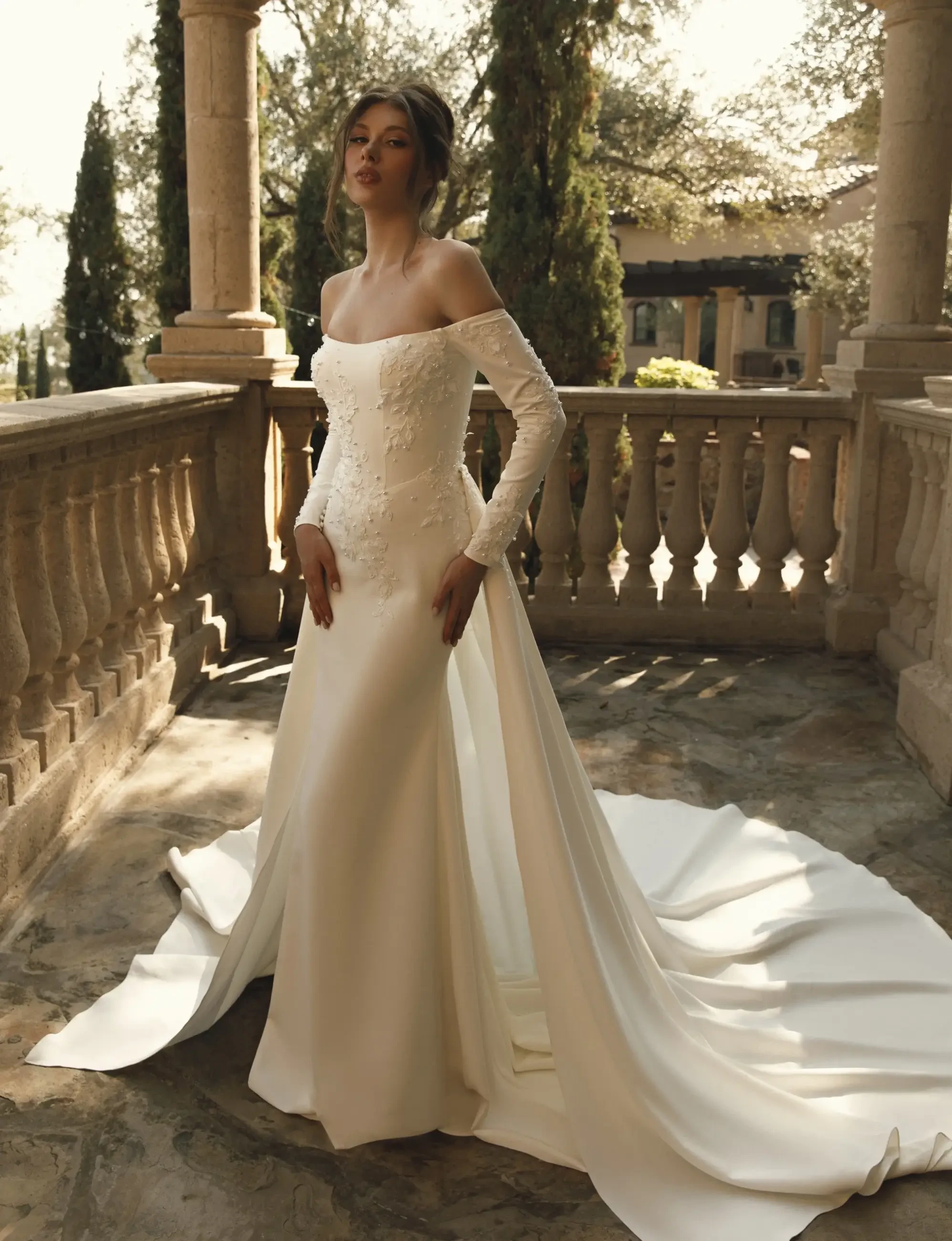 A model in an elegant off-the-shoulder white wedding gown with long sleeves, standing on a stone balcony surrounded by greenery. The gown features intricate detailing and a flowing train.