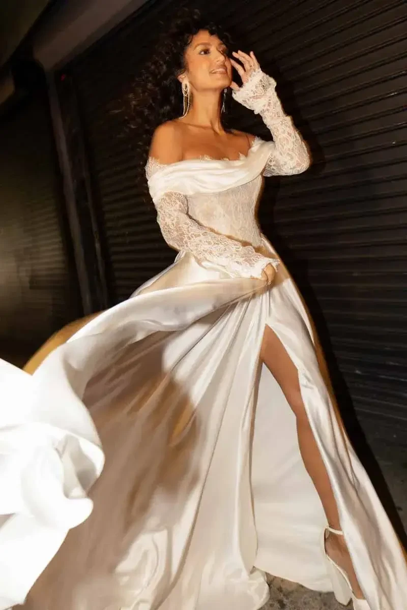 A woman in a flowing white gown with lace sleeves and a high slit, walking confidently down a street.