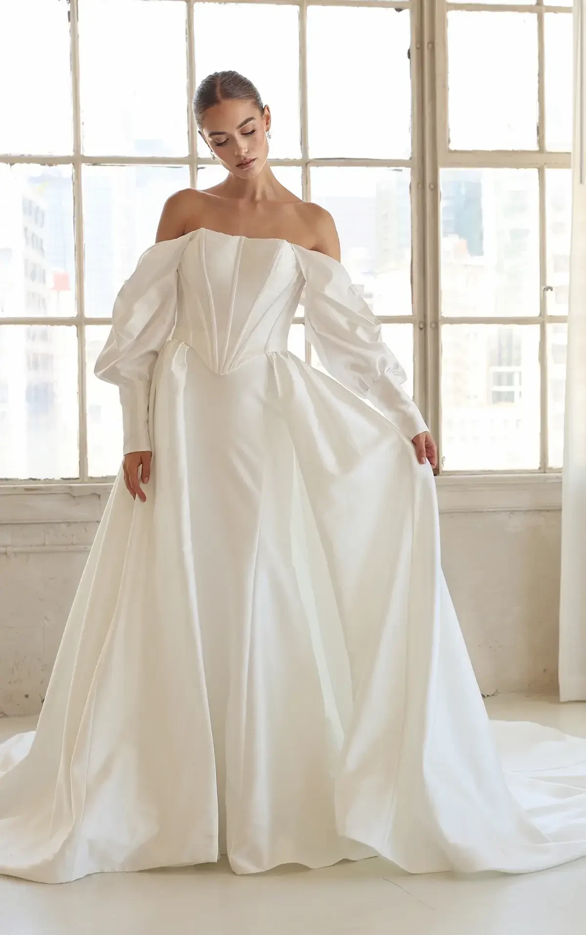 Model is wearing a strapless silk column gown featuring detachable long blouson sleeves and a detachable silk overskirt