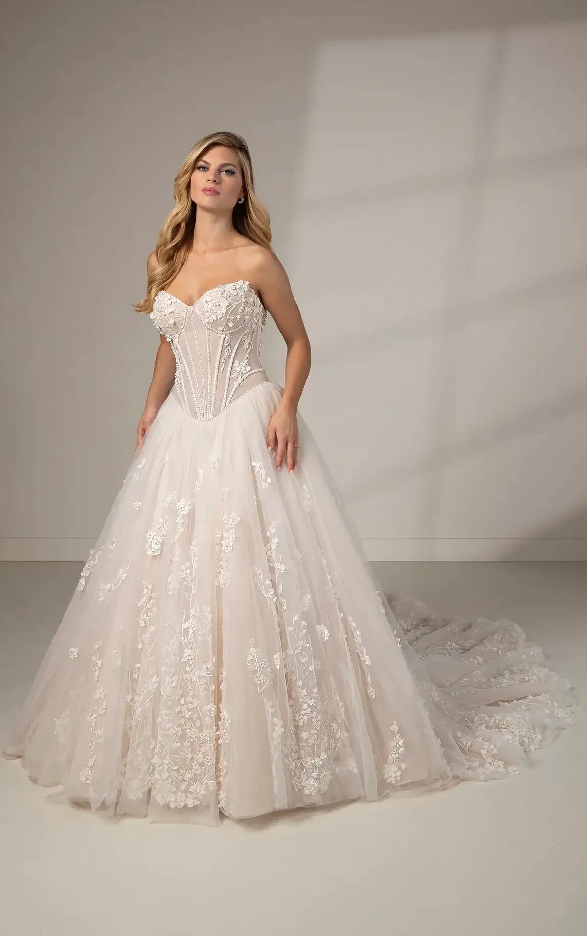 Model is wearing a strapless sweetheart ball gown with a sheer corset bodice, pearl-beaded detailing, 3D floral appliqués, lace-up back, and a long tulle skirt with a romantic train.