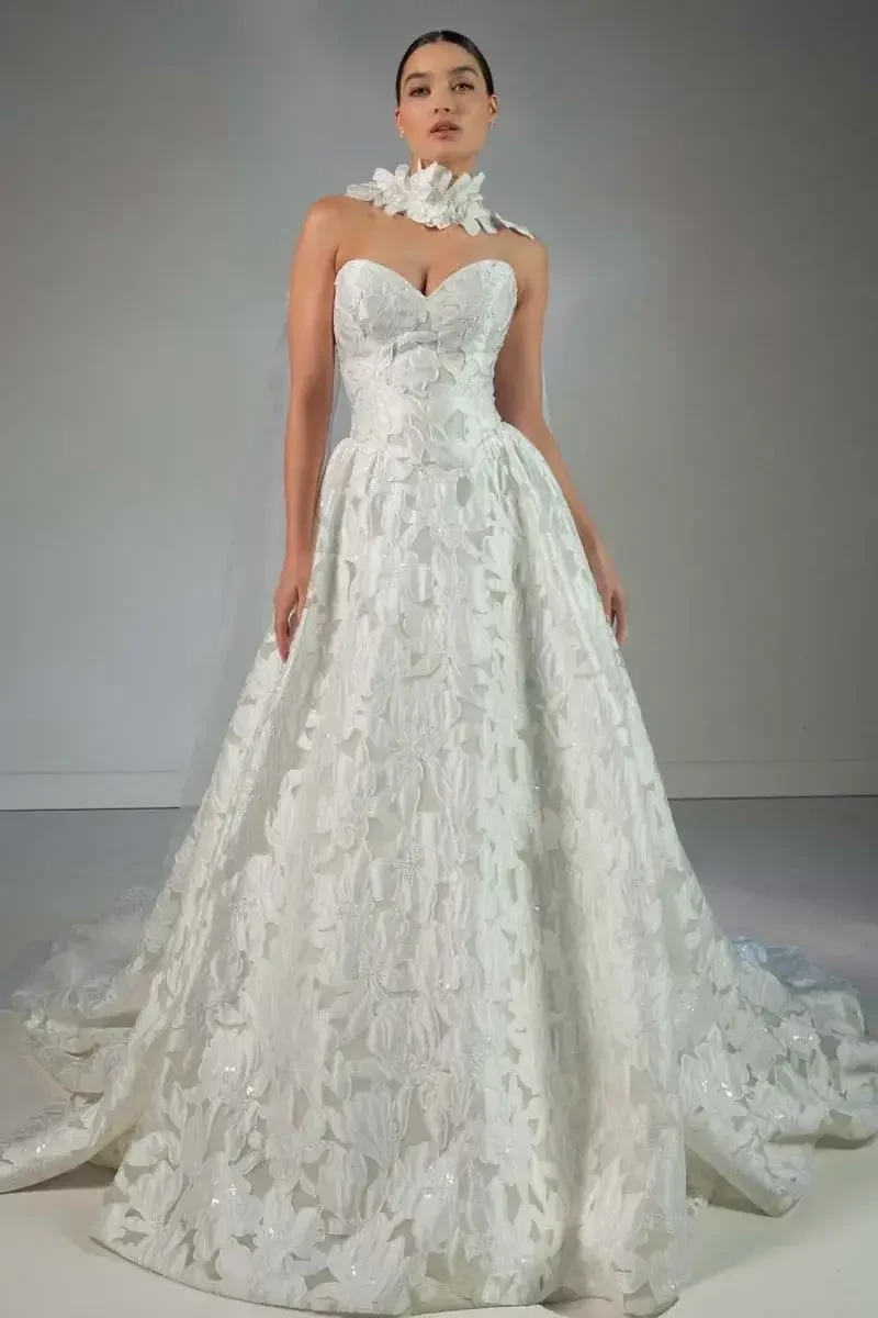A model wearing an elegant white gown with a floral design, posing against a neutral background.