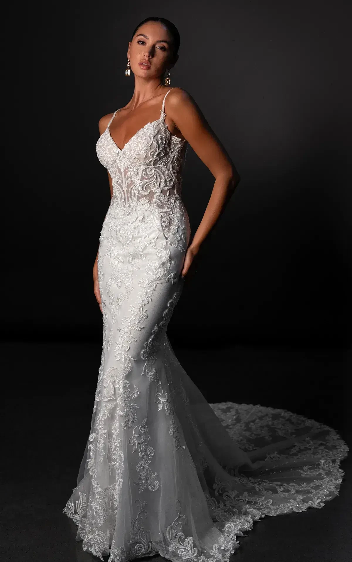 A woman in an elegant white bridal gown with intricate lace details, standing confidently against a dark background.