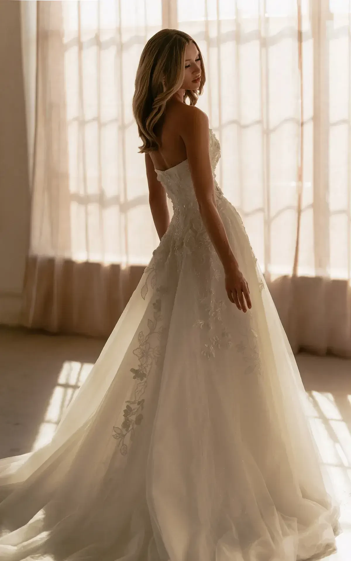 A woman stands in soft, natural light wearing a strapless, A-line wedding dress with floral lace. Her back is to the camera, evoking a serene, romantic mood.