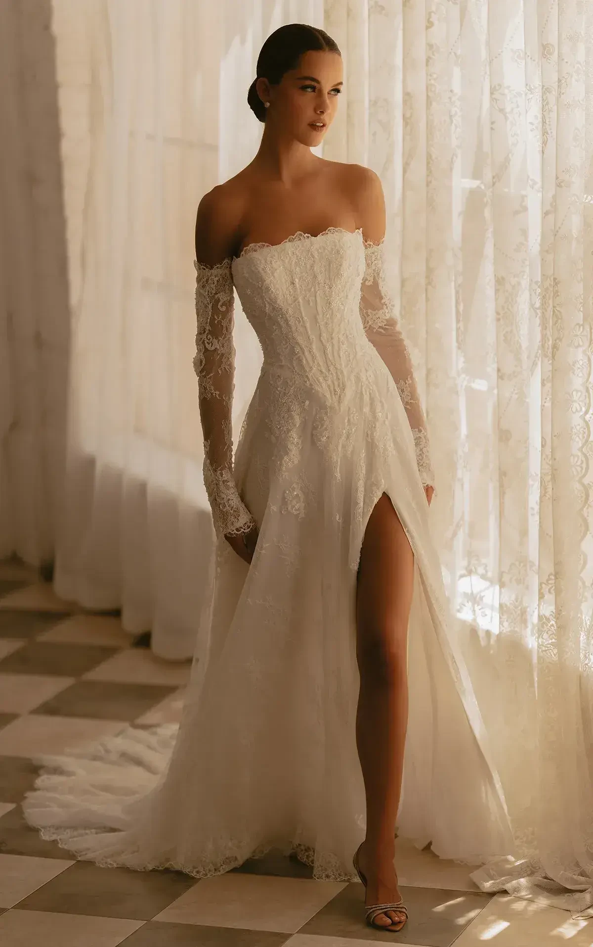 A woman in a white off-the-shoulder wedding dress with long lace sleeves and a thigh-high slit, standing by a window with light filtering through sheer curtains.