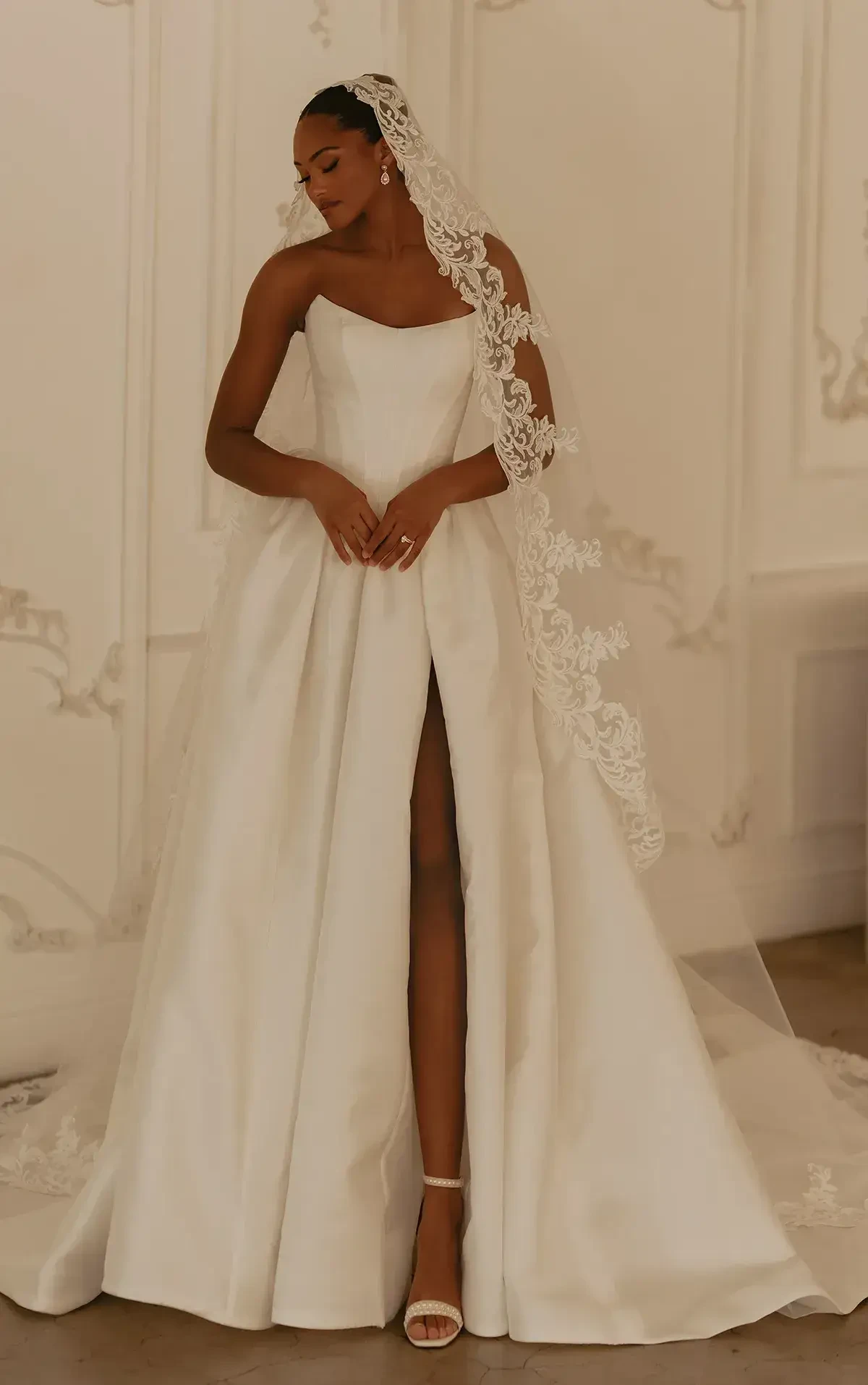 A woman in a white wedding dress with a high slit and lace detailing, wearing a long veil, stands elegantly in front of an ornate backdrop.