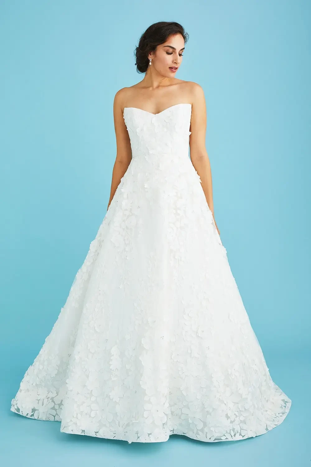 A woman in a strapless white wedding gown with floral applique stands against a blue background, exuding elegance and poise.