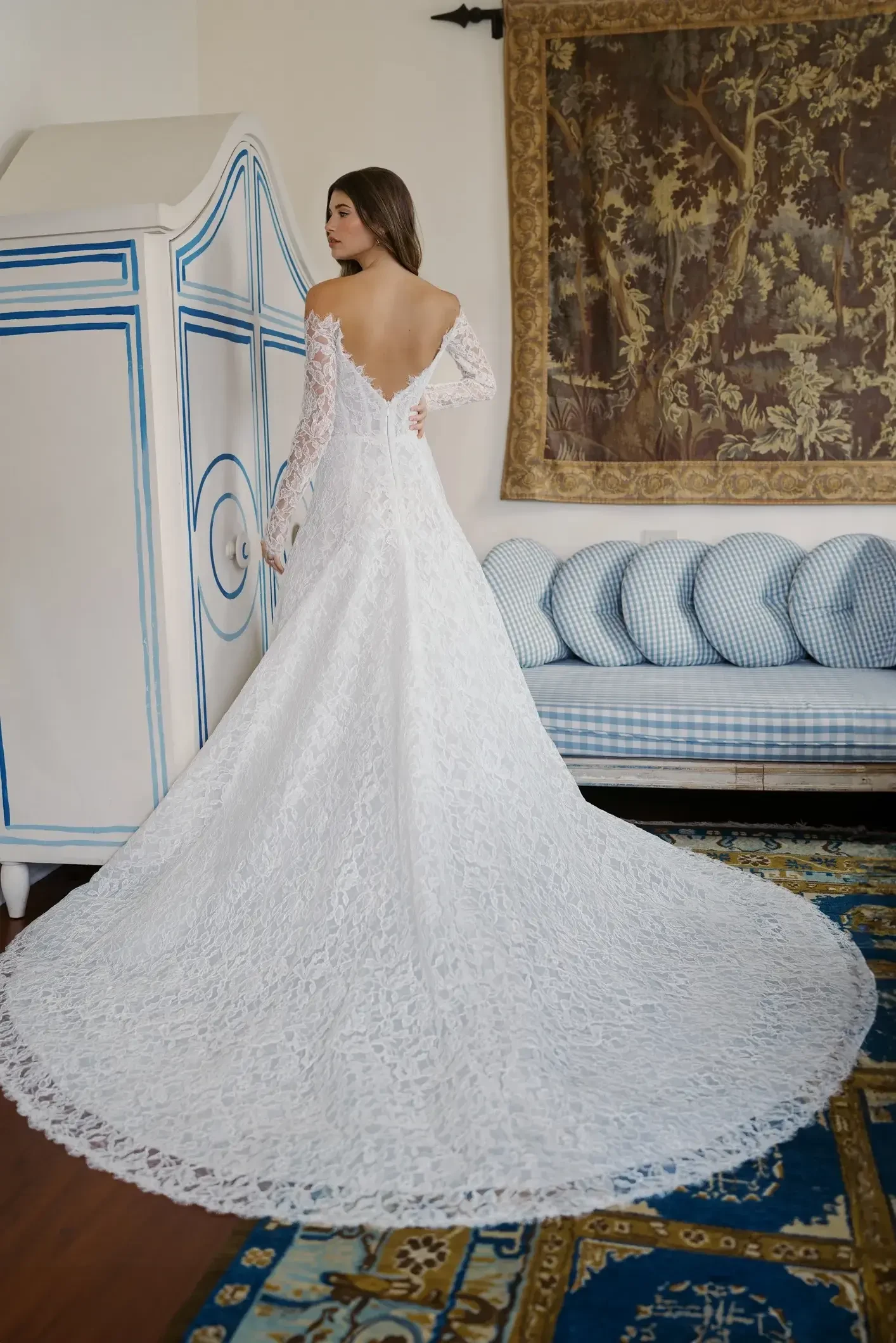 Model is wearing a long-sleeved modified A-line gown with corded lace, a portrait neckline, and an extended dramatic train with eyelash trim.