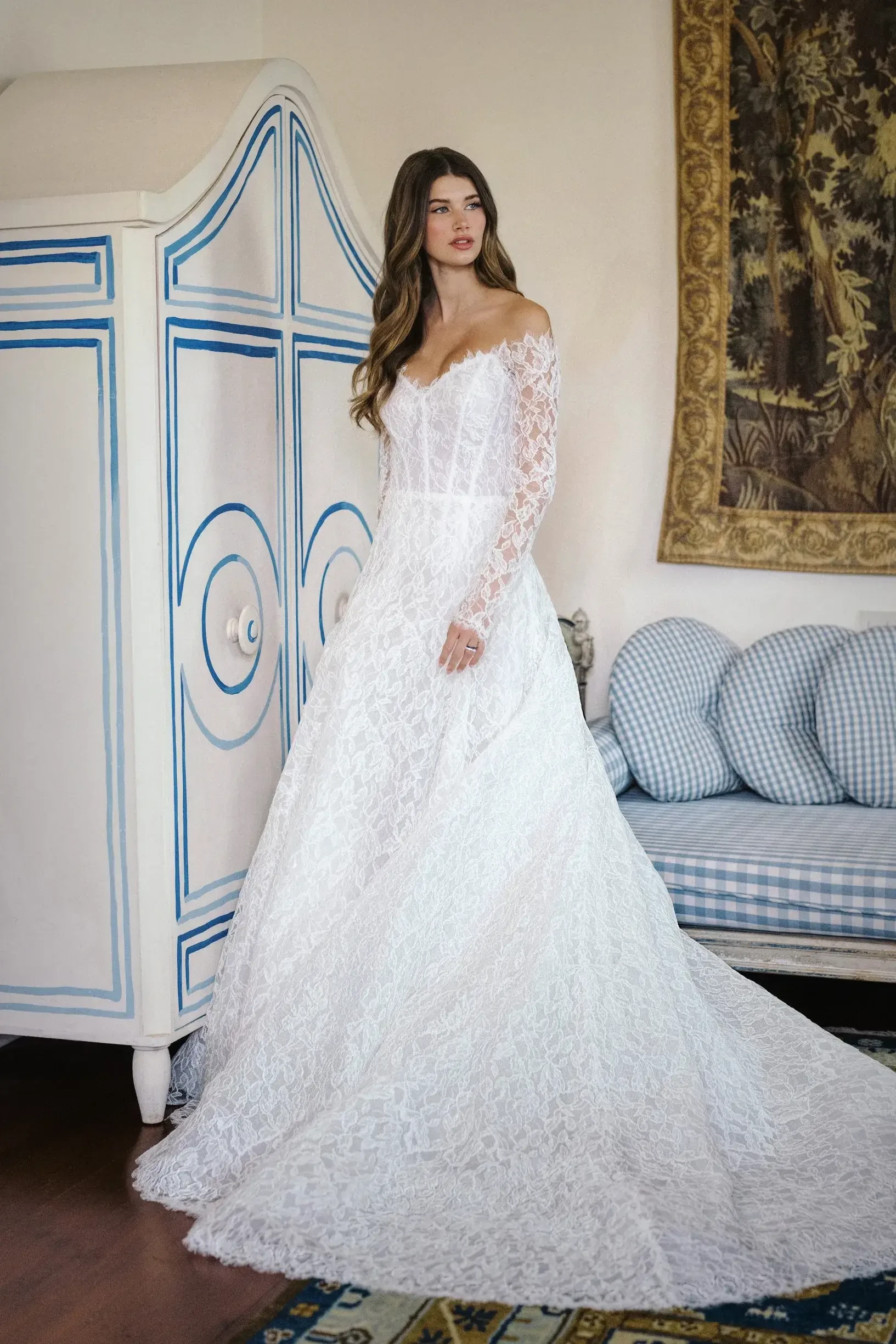 Model is wearing a long-sleeved portrait neckline gown in corded lace with a modified A-line silhouette, eyelash trim, and a dramatic extended train.